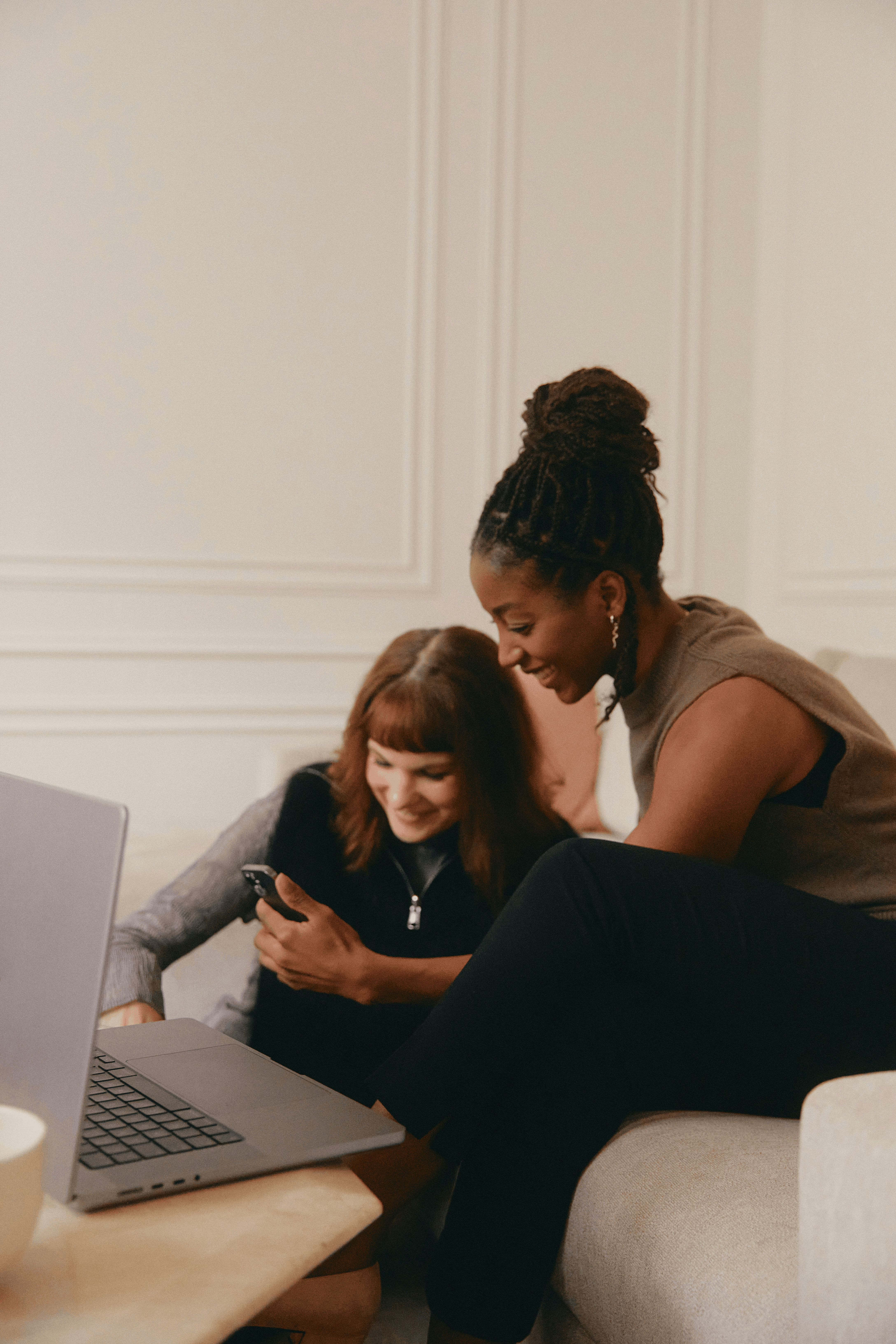 Two people looking at a smartphone beside a laptop in a bright living room